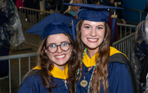 two graduating students smile at the camera. they wear caps and gowns regalia.